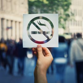 Hand holding a paper sheet with no smoking sign over a crowded street public place background. Forbidden area zone, restrictive symbol stop smoke.